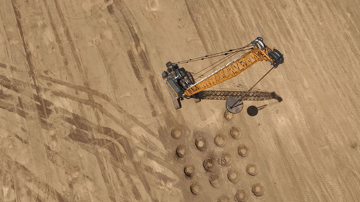Aerial view of a dynamic compaction crane lifting a heavy steel weight above patterned soil impact craters on a construction site.