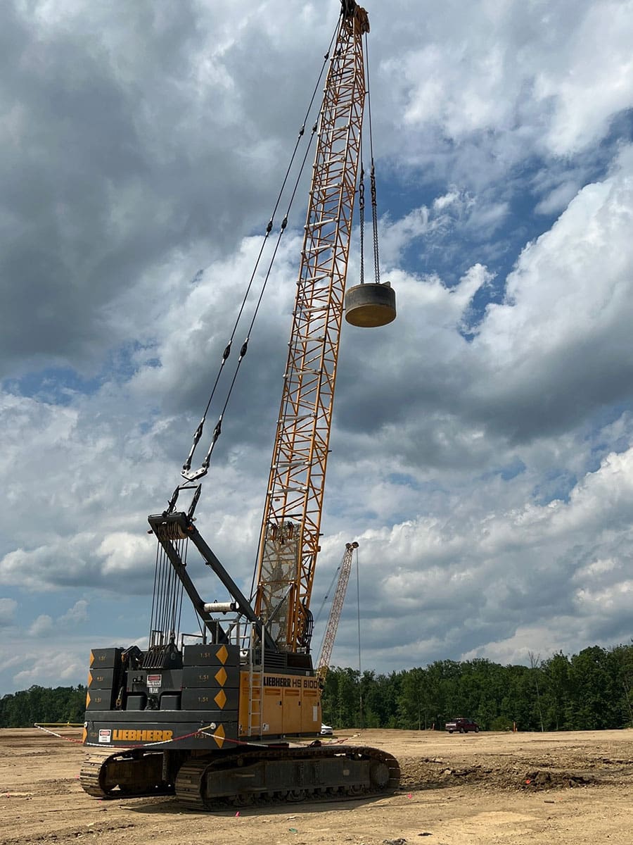 Crawler crane lifting a heavy steel weight used for dynamic compaction to strengthen soil on a large construction site under a cloudy sky.