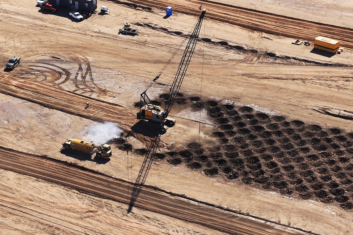 Aerial view of a dynamic compaction crane dropping a heavy weight to densify soil, creating a grid of impact craters on a construction site.