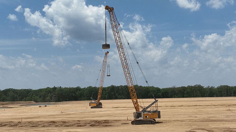 Two crawler cranes dropping heavy tampers for dynamic compaction to densify soil at a large construction site.