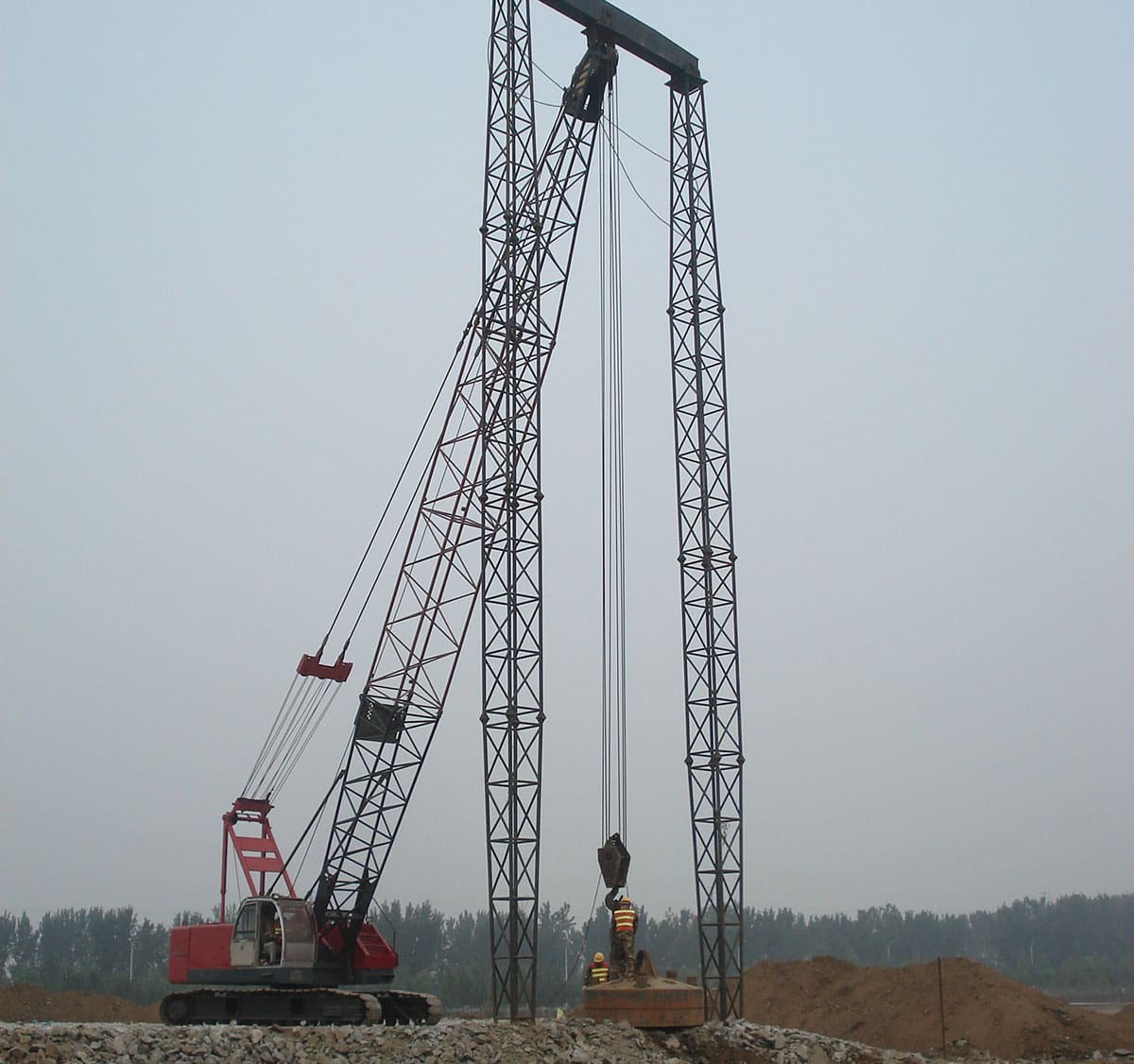 Crawler crane lifting a heavy compaction weight between tall guide frames with workers assisting below.