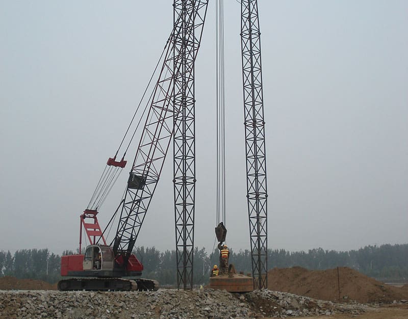 crane-guided-dynamic-compaction-operation-densification Crawler crane lifting and positioning a heavy tamper weight between guide towers during dynamic compaction soil densification.