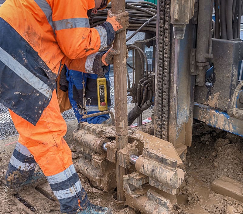 Worker operating geotechnical drilling equipment to test soil conditions on a muddy construction site during ground investigation.