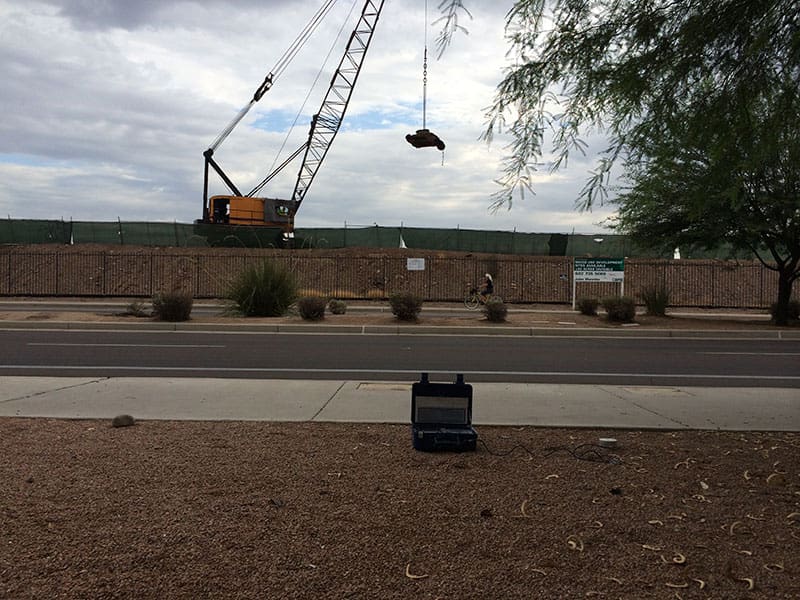 Crawler crane lifting tamper weight for dynamic compaction while vibration monitoring equipment records ground movement near roadway.