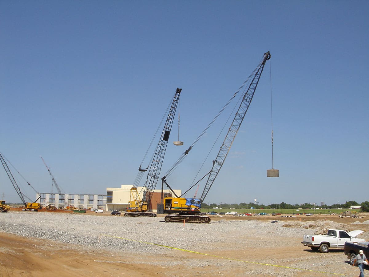 multiple-cranes-dynamic-compaction-site-densification Two crawler cranes lifting heavy tamper weights for dynamic compaction on a large industrial construction site with buildings in the background.