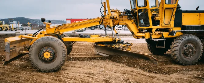 Yellow motor grader leveling soil at a construction site, preparing the ground for infrastructure development under overcast skies.