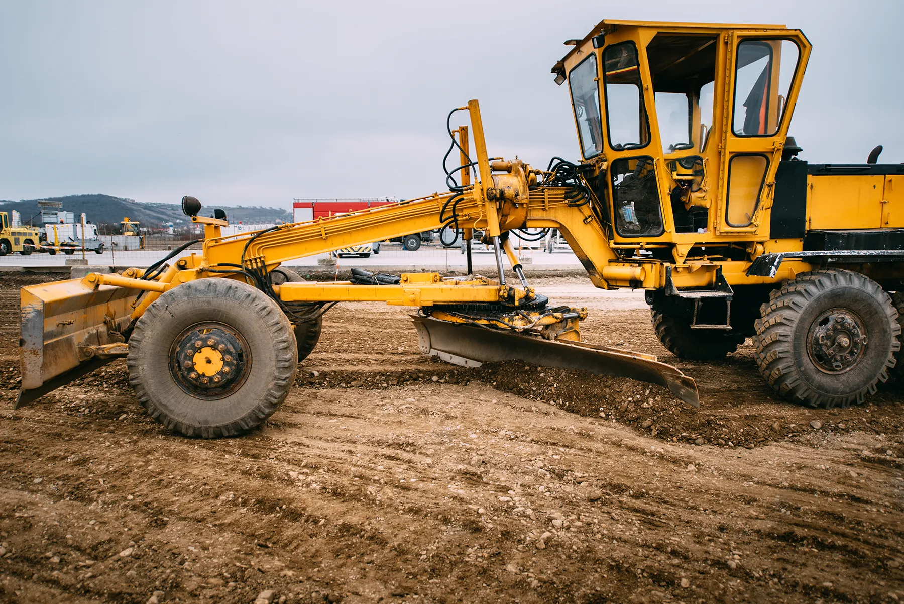Yellow motor grader leveling soil at a construction site, preparing the ground for infrastructure development under overcast skies.