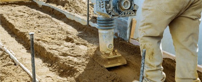 Worker using a plate compactor to compact sandy soil along a foundation trench during site preparation for construction.