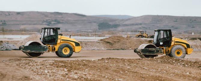 Two soil compaction rollers compacting dirt across a large construction site with earthmoving equipment and hills in the background.
