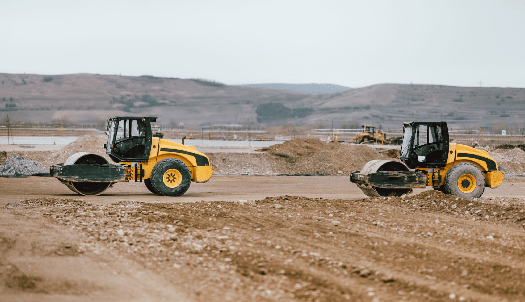 Two soil compaction rollers compacting dirt across a large construction site with earthmoving equipment and hills in the background.