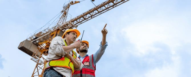 Two construction workers in safety gear communicating near a large crane on a job site, emphasizing coordination and safety in crane operations.