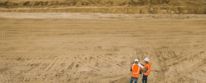 Two construction workers in high-visibility vests and hard hats stand on a large sandy construction site, reviewing plans while heavy machinery operates in the background.