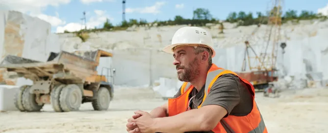 Tired worker of marble quarry sitting in front of camera by workplace against dump truck and working bulldozer with crane