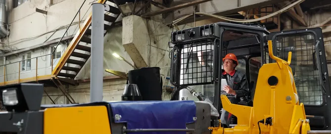 Young contemporary female worker in workwear and safety helmet operating huge construction machine during work