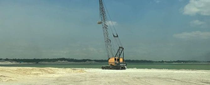 Crawler crane performing dynamic compaction on a sandy coastal construction site near water during ground improvement work.