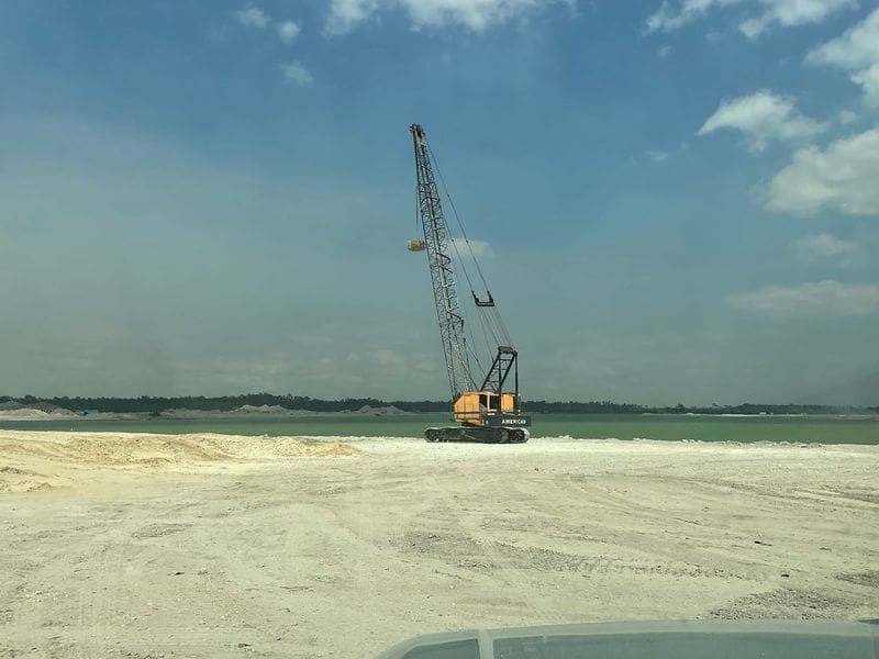 Crawler crane performing dynamic compaction on a sandy coastal construction site near water during ground improvement work.