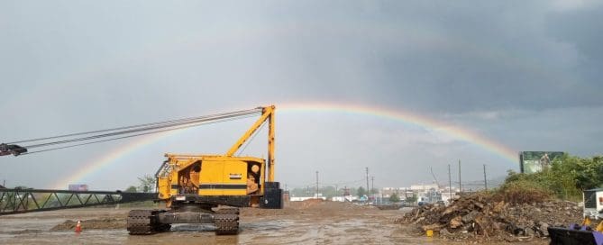 A yellow crawler crane on a muddy construction site with a double rainbow arching across a grey sky in the background.