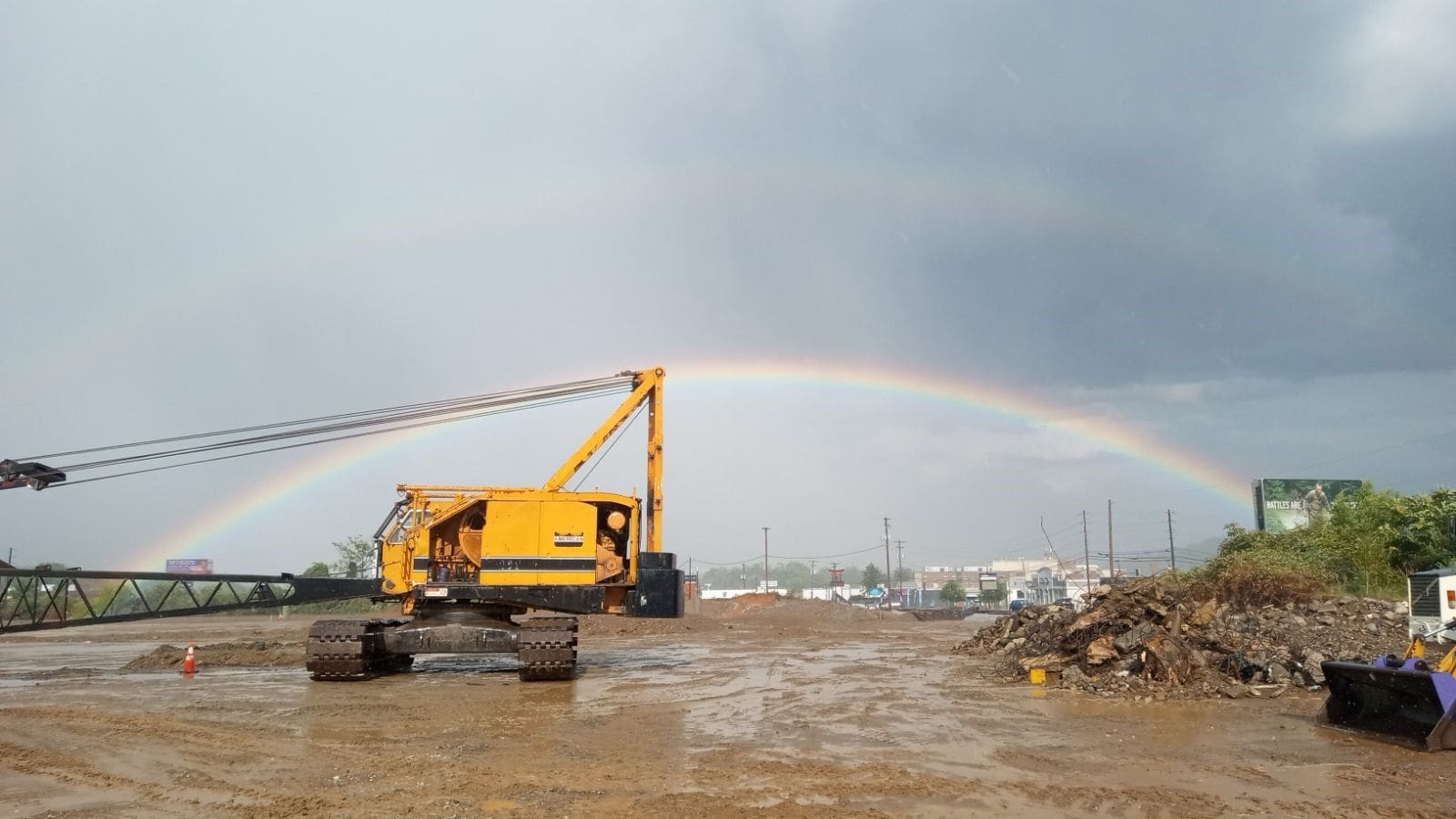 A yellow crawler crane on a muddy construction site with a double rainbow arching across a grey sky in the background.