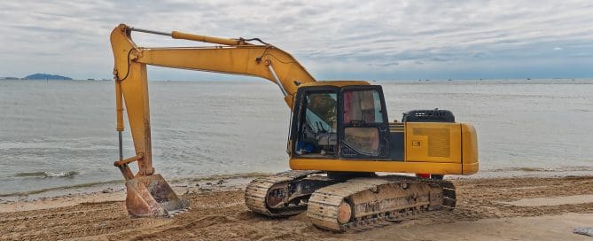 Yellow excavator parked on a sandy beach near the ocean, with tire tracks in the sand and overcast skies above.