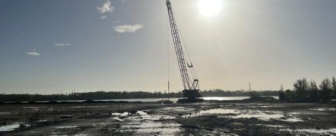 A wide-angle silhouette of a lattice boom crane on a flat, muddy construction site with the bright sun directly above it.