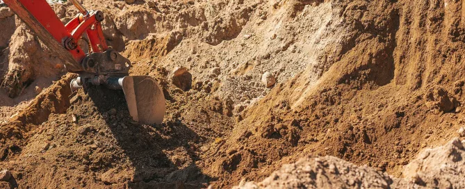 A close-up view of an orange excavator bucket digging into a mound of brown earth.