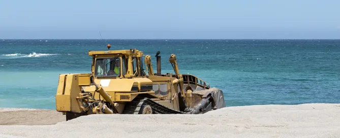 Bulldozer grading sand along a coastal construction site, illustrating soil stabilization and ground improvement work near shoreline structures.