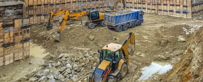 Excavators and trucks preparing a construction site near existing structures, illustrating soil excavation and ground improvement work in an urban environment.