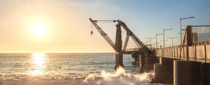 A silhouette of a large industrial crane situated on a pier at sunset, with waves crashing against the support pillars and a golden light reflecting off the ocean.
