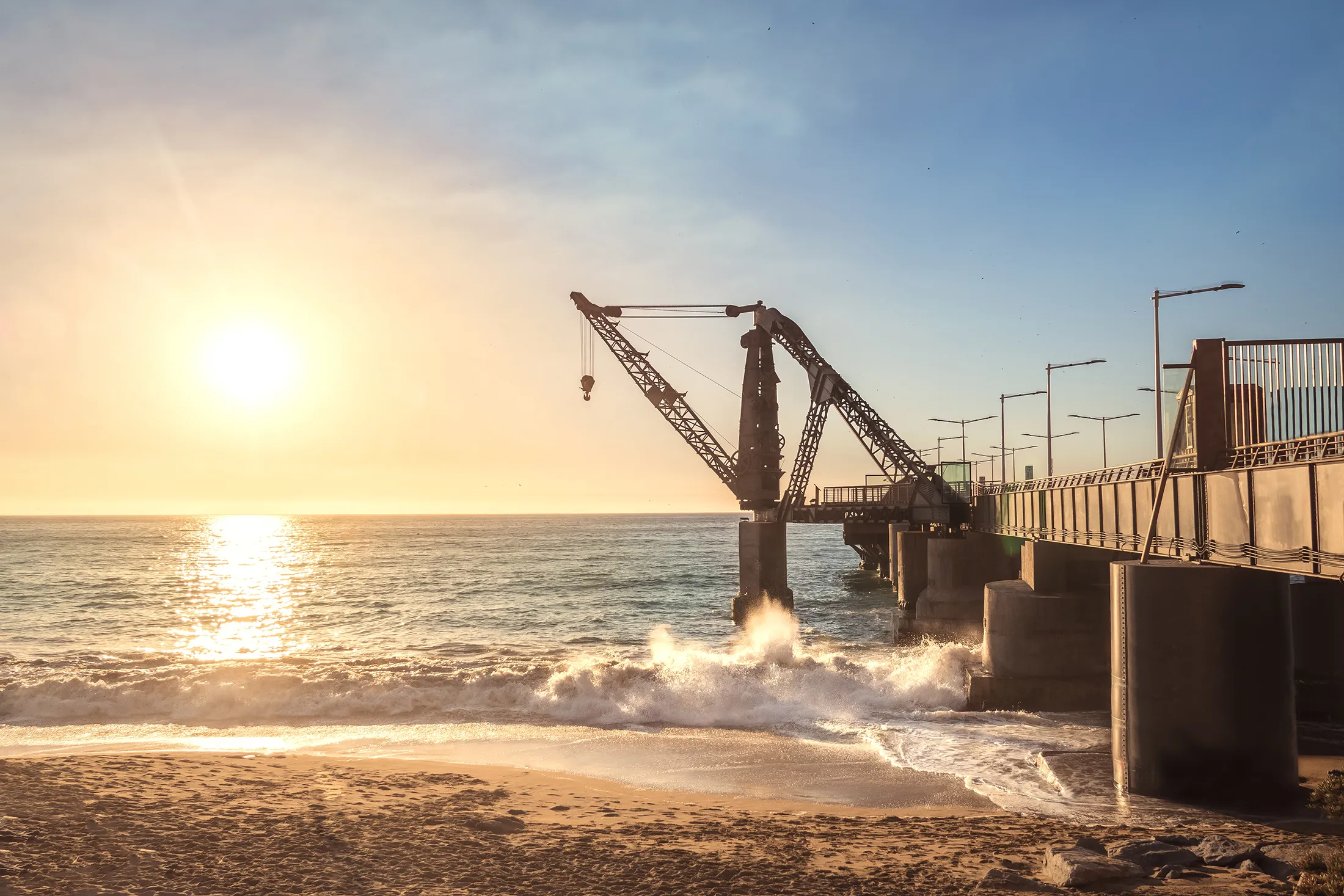 A silhouette of a large industrial crane situated on a pier at sunset, with waves crashing against the support pillars and a golden light reflecting off the ocean.