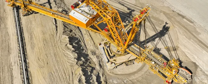 An overhead aerial view of a massive yellow bucket-wheel excavator working in a deep open-pit mine, showing the intricate lattice structure and the conveyor belt system.
