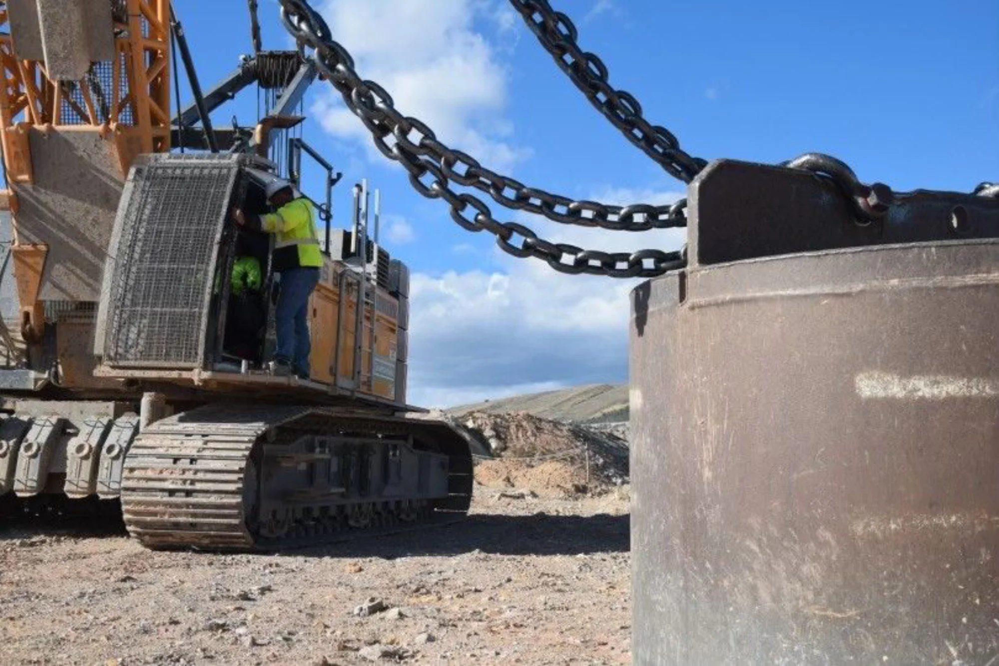 A close-up of a construction worker in a high-visibility vest standing on the tracks of a large crane, with heavy metal chains connected to a massive steel weight in the foreground.