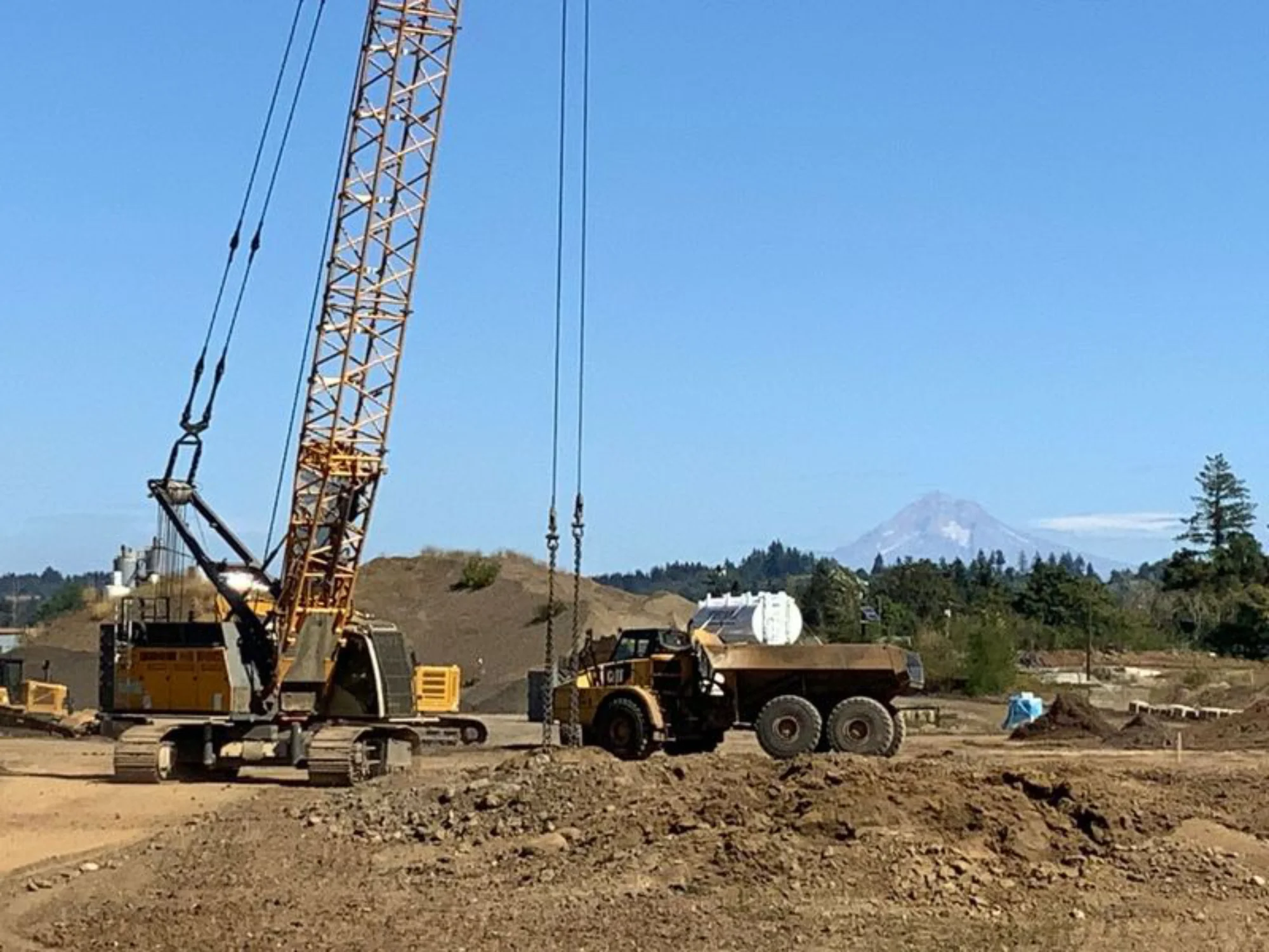 A large crawler crane with a lattice boom standing on a dirt construction site next to a yellow dump truck, with a snow-capped mountain visible in the distant background.