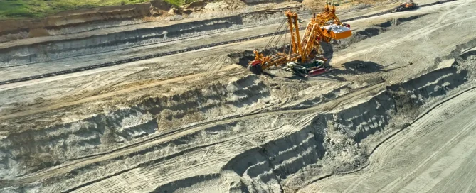 An aerial view of a massive yellow bucket-wheel excavator operating in a large open-pit mine with terraced earth walls and a forest in the background.