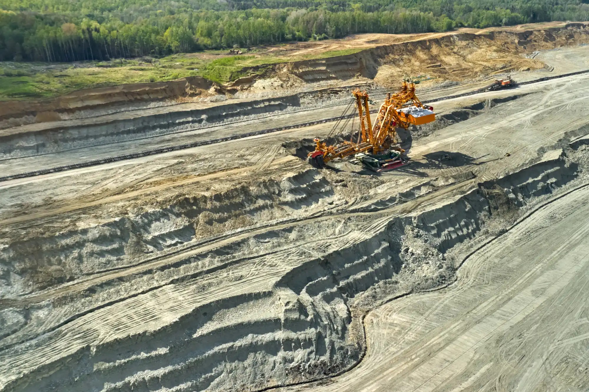 An aerial view of a massive yellow bucket-wheel excavator operating in a large open-pit mine with terraced earth walls and a forest in the background.