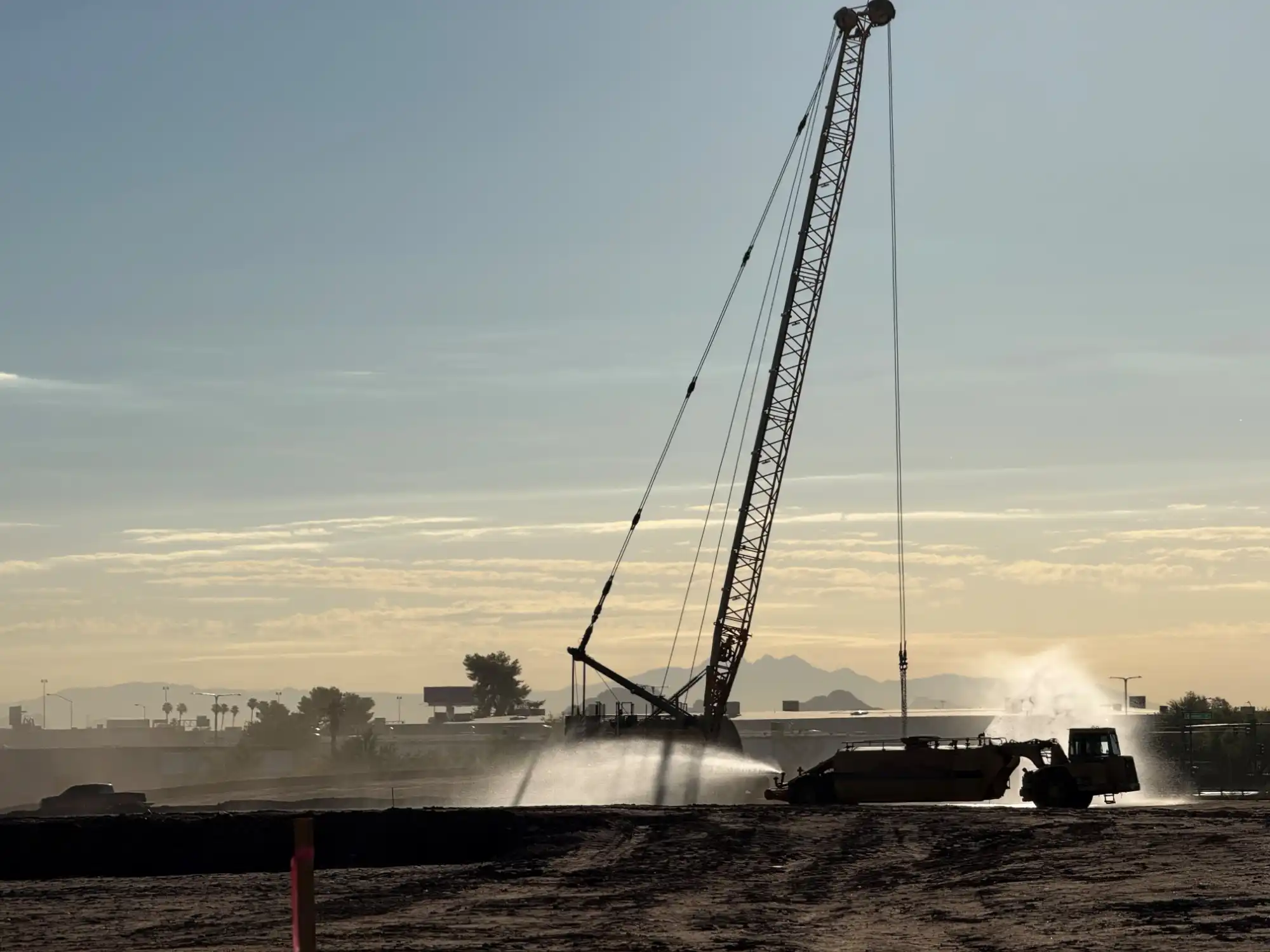 crawler-crane-dynamic-compaction-dusk-site-densification. Crawler crane operating over a dynamic compaction site at dusk with a water truck suppressing dust nearby