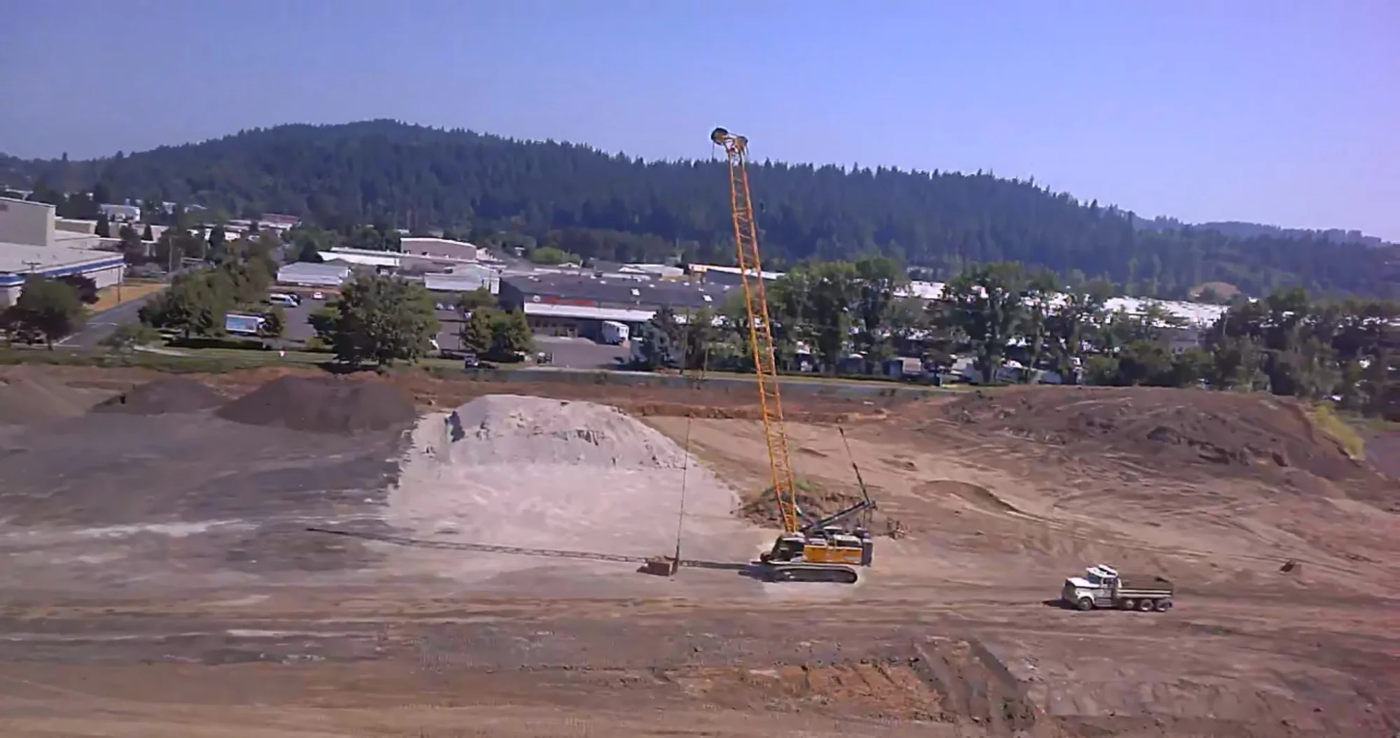 dynamic-compaction-crane-soil-stockpile-densification. Dynamic compaction crane lifting a tamping weight above a soil stockpile at an industrial site