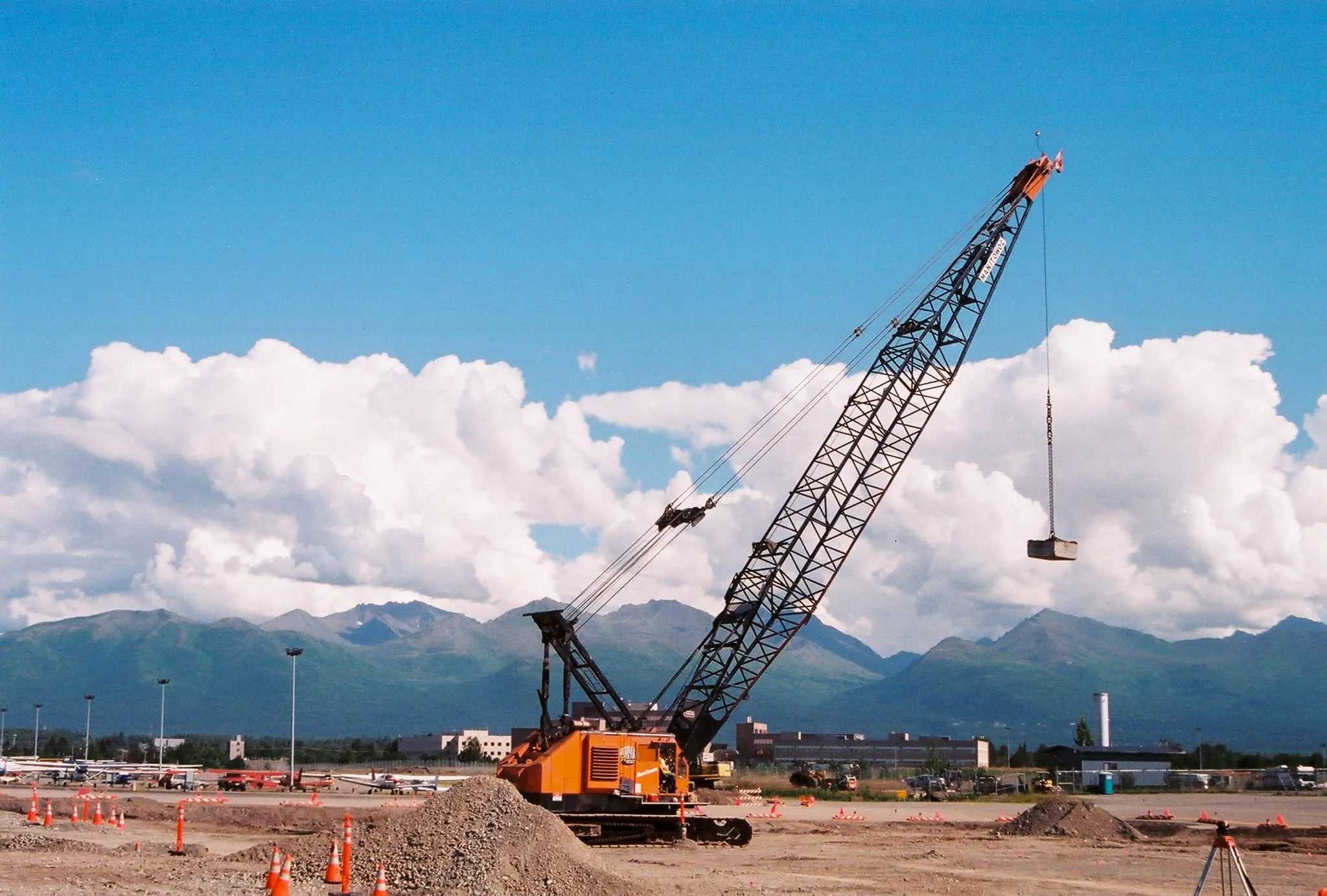 manitowoc-crane-airfield-compaction-anchorage-densification. Orange Manitowoc crawler crane with a drop weight raised over an Anchorage airfield site, Chugach peaks behind