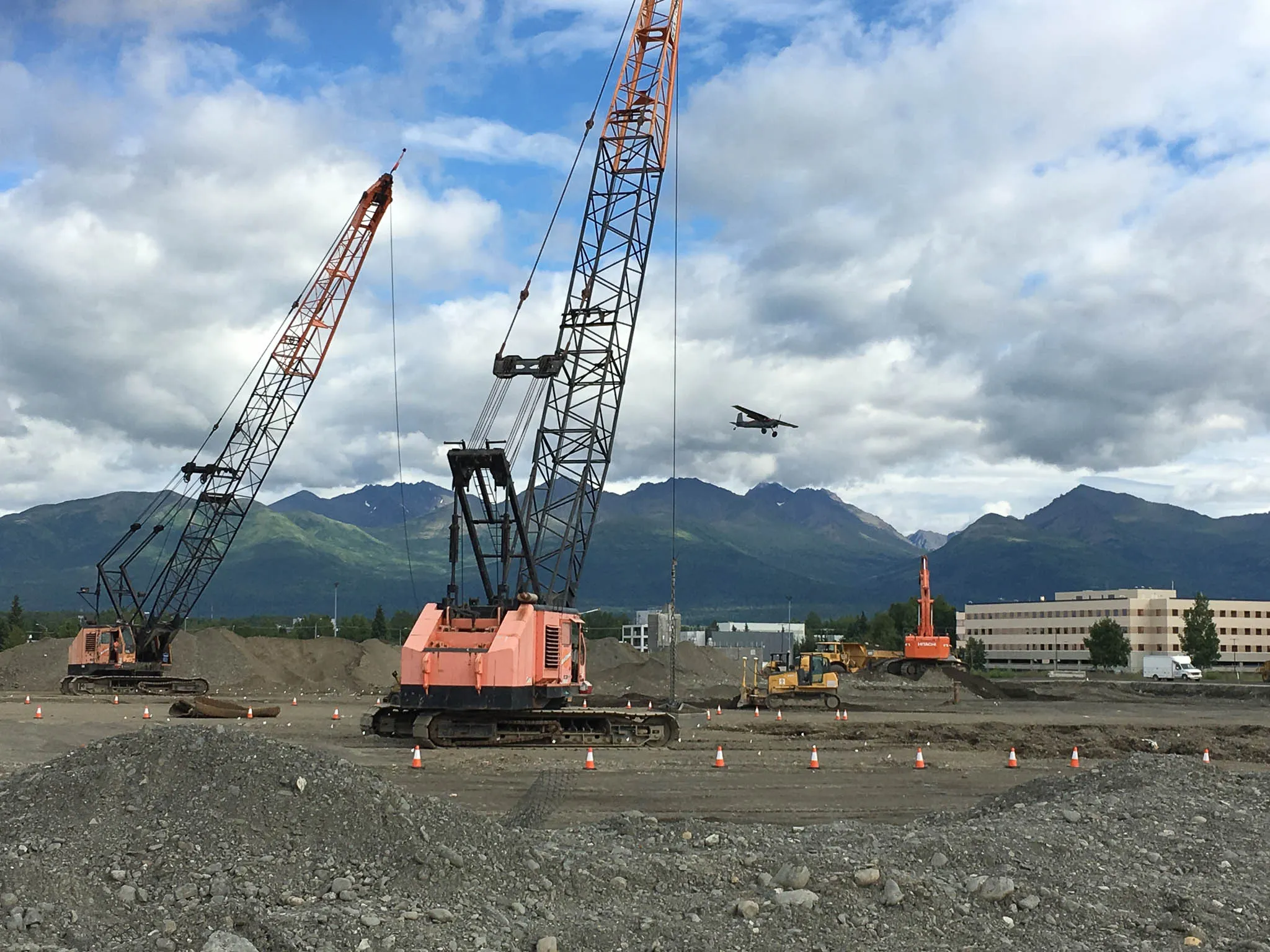 two-crawler-cranes-airfield-site-anchorage-densification. Two orange crawler cranes on an Anchorage ground improvement site as a small plane passes overhead