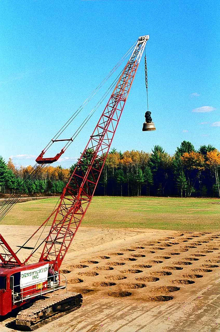 red-crawler-crane-dynamic-compaction-densification. Red crawler crane suspends a steel tamper above a completed dynamic compaction crater grid