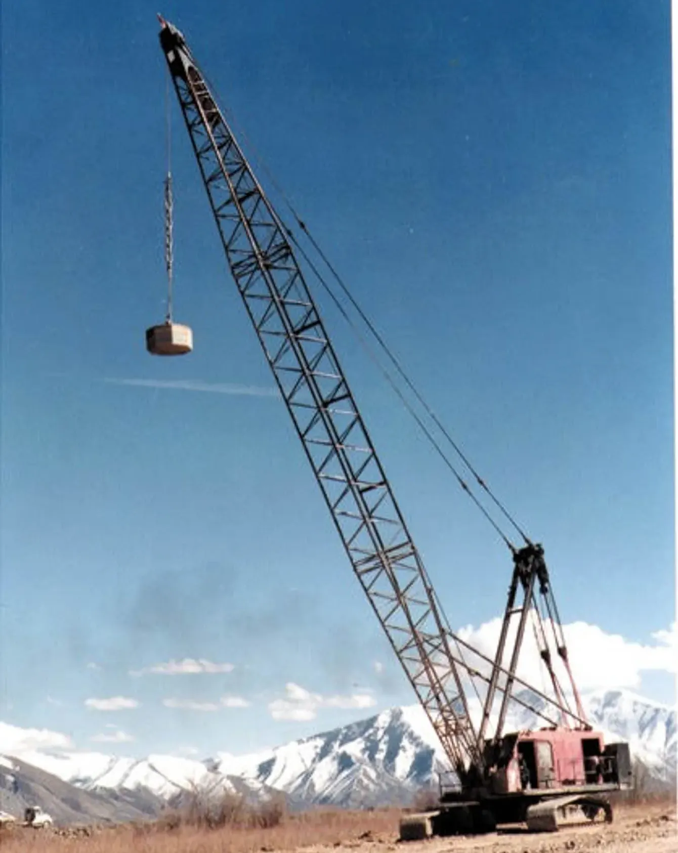 crane-tamper-snowy-mountains-provo-utah-densification. Red lattice-boom crawler crane holding a tamper high above a Provo, Utah site with snow-capped peaks behind