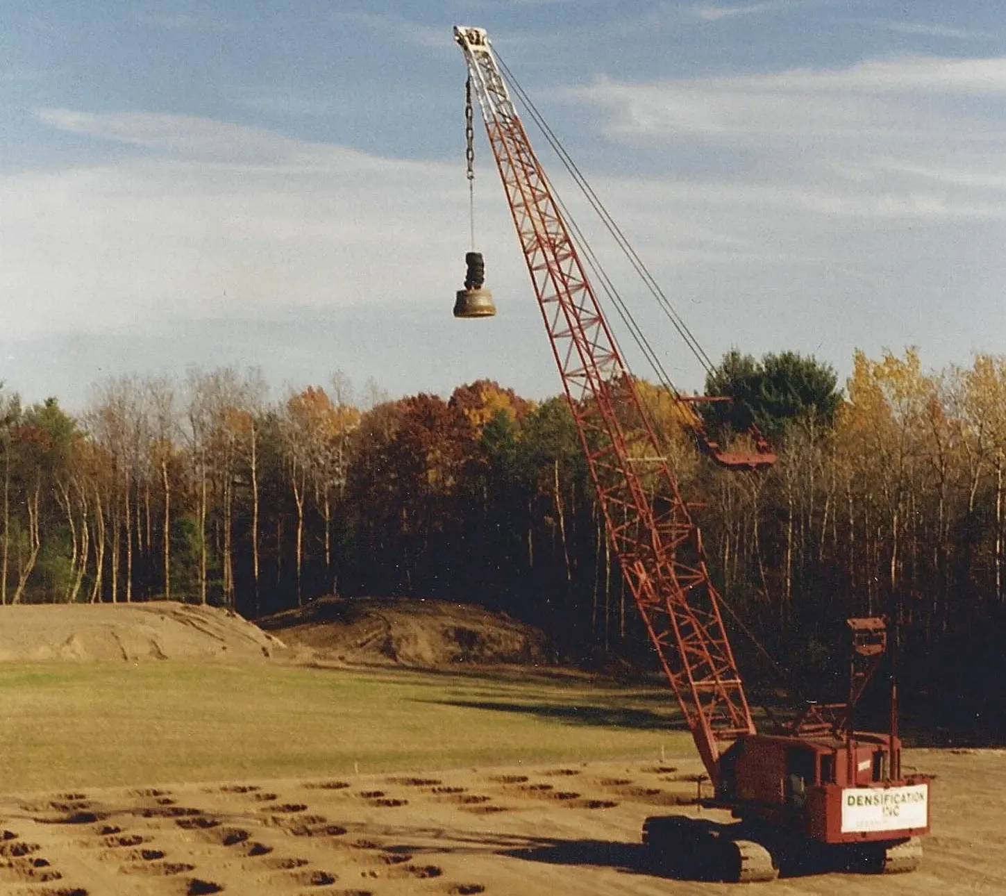 densification-inc-crane-autumn-site-saratoga-springs-densification. Red Densification Inc crawler crane dropping a tamper across a sand site framed by autumn foliage