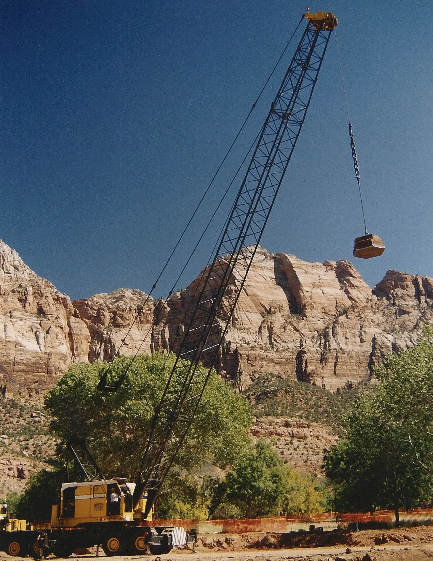 crane-lifting-tamper-zion-canyon-utah-densification. Yellow truck-mounted lattice crane lifting a tamper below the red sandstone cliffs of Zion, Utah